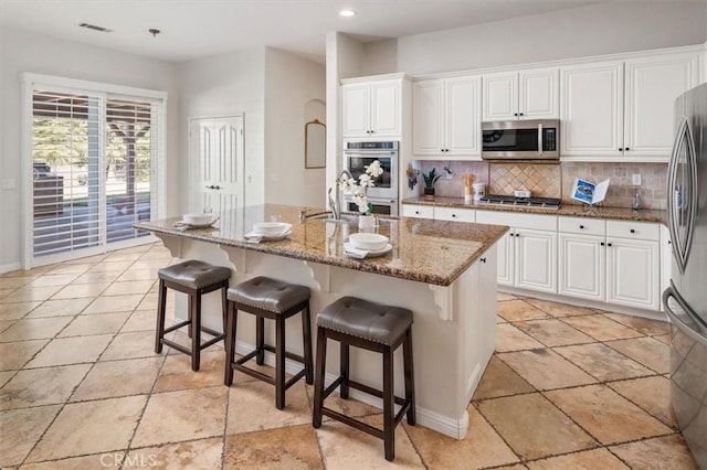 kitchen featuring tasteful backsplash, a center island with sink, dark stone counters, appliances with stainless steel finishes, and white cabinetry