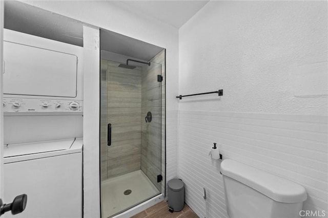 bathroom with a wainscoted wall, stacked washer / drying machine, toilet, a shower stall, and wood finished floors