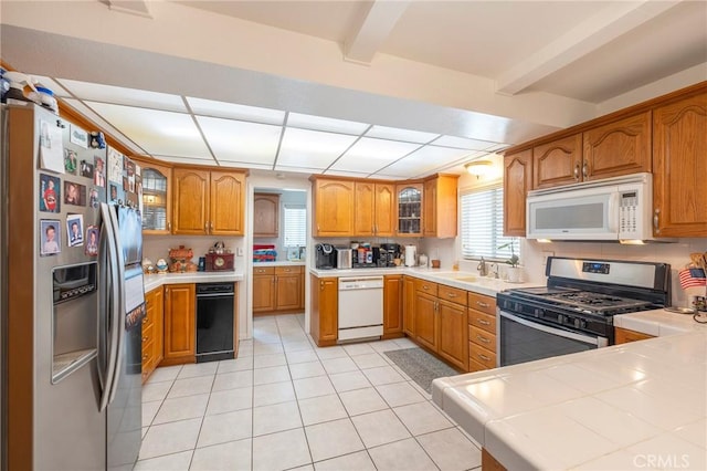 kitchen featuring light tile patterned flooring, appliances with stainless steel finishes, brown cabinets, beamed ceiling, and glass insert cabinets