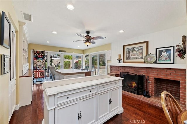 kitchen with an island with sink, dark wood-style flooring, light countertops, a brick fireplace, and white cabinetry