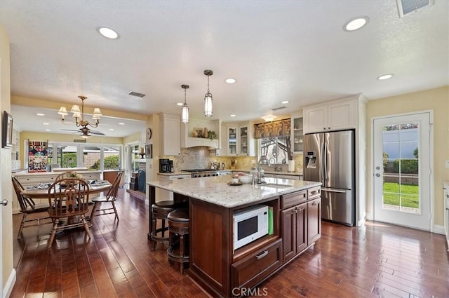 kitchen with hanging light fixtures, white microwave, glass insert cabinets, an island with sink, and stainless steel fridge