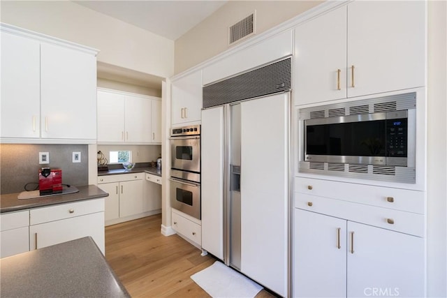 kitchen with white cabinets, light wood finished floors, dark countertops, visible vents, and built in appliances