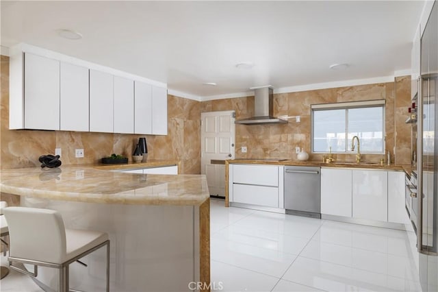 kitchen featuring light countertops, white cabinetry, wall chimney range hood, dishwasher, and a peninsula