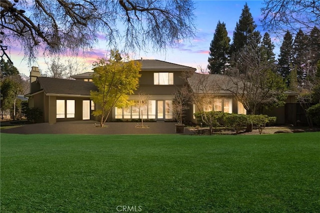 back of house at dusk featuring a tiled roof, a lawn, a chimney, and stucco siding