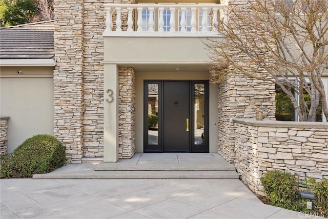 view of exterior entry with stone siding, a balcony, and stucco siding