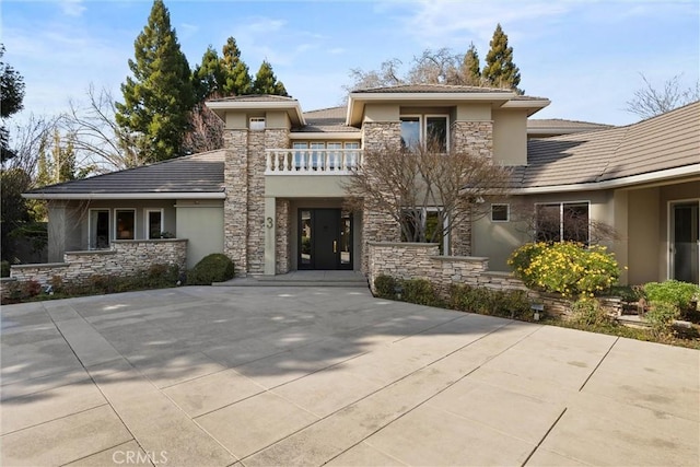 prairie-style home with stone siding, a tile roof, a balcony, and stucco siding