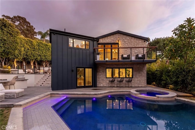 back of house with a balcony, a pool with connected hot tub, stairs, stone siding, and board and batten siding