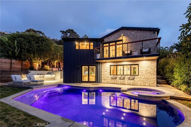 back of house featuring a pool with connected hot tub, board and batten siding, a patio area, fence, and a balcony