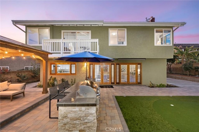 back of property at dusk with a patio, a balcony, exterior kitchen, french doors, and stucco siding