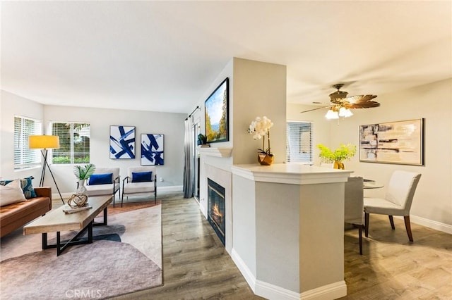 living area featuring ceiling fan, baseboards, wood finished floors, and a glass covered fireplace