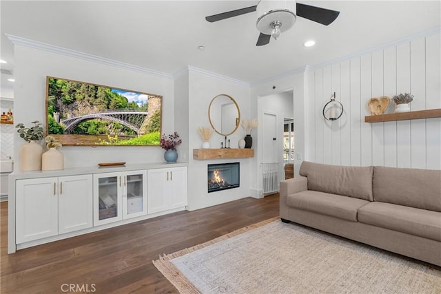 living area with dark wood-type flooring, ornamental molding, a warm lit fireplace, recessed lighting, and ceiling fan