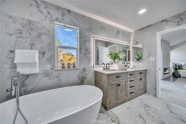 full bath with double vanity, a soaking tub, marble finish floor, stone wall, and a sink