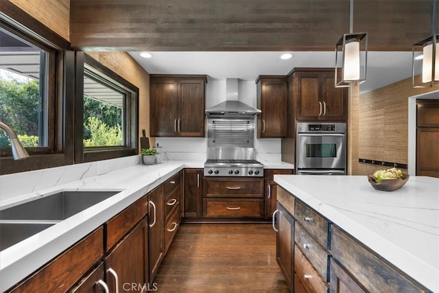 kitchen featuring light stone counters, stainless steel appliances, a sink, wall chimney range hood, and pendant lighting