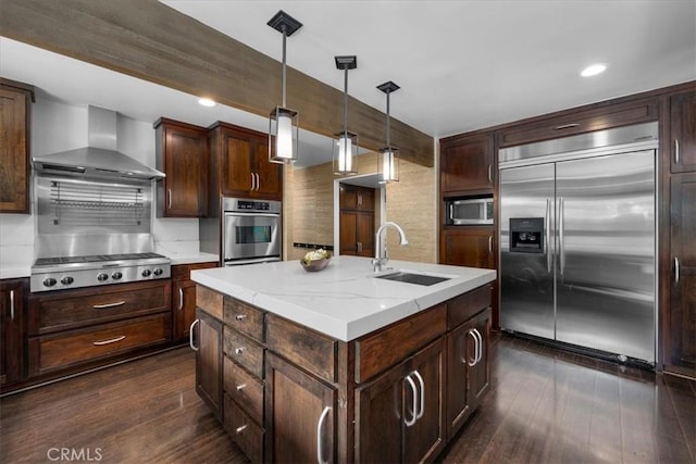 kitchen featuring decorative light fixtures, appliances with stainless steel finishes, a kitchen island with sink, a sink, and wall chimney exhaust hood