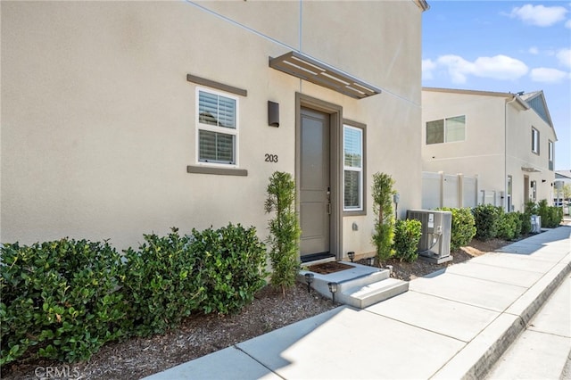 view of exterior entry with cooling unit, fence, and stucco siding