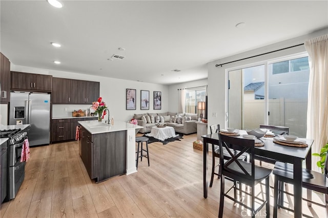 kitchen with dark brown cabinetry, a center island with sink, open floor plan, stainless steel appliances, and light countertops