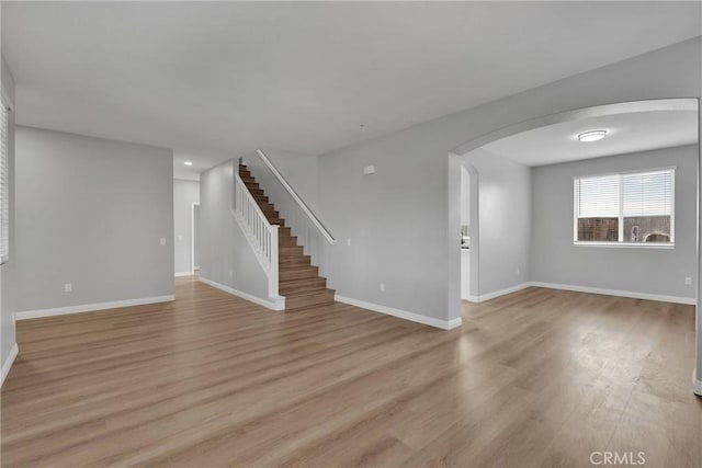 empty room with arched walkways, stairway, baseboards, and light wood-style floors