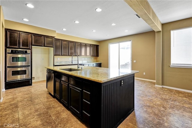 kitchen with baseboards, appliances with stainless steel finishes, light stone counters, a kitchen island with sink, and a sink