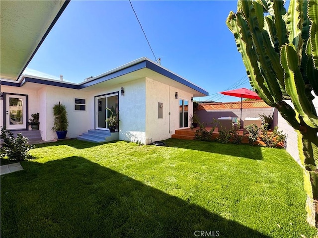 rear view of house featuring entry steps, fence, a lawn, and stucco siding