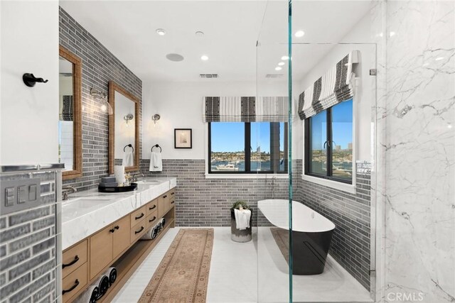 full bathroom featuring double vanity, visible vents, tile walls, a freestanding tub, and a sink