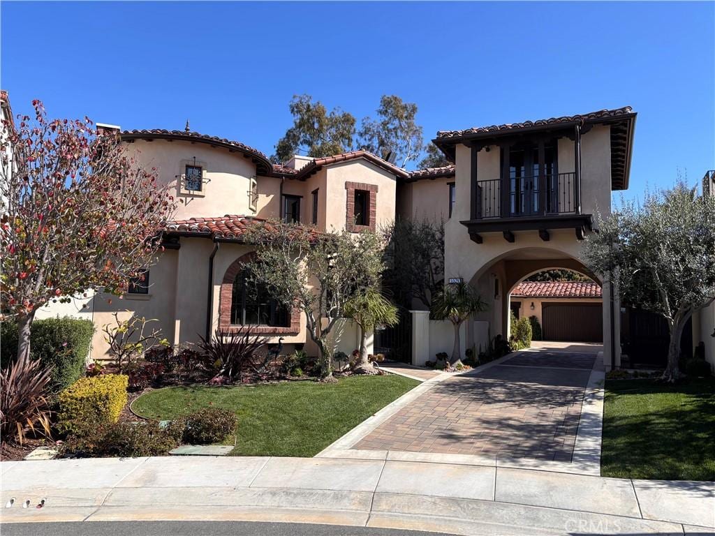 mediterranean / spanish house with stucco siding, decorative driveway, a front yard, a balcony, and a tiled roof