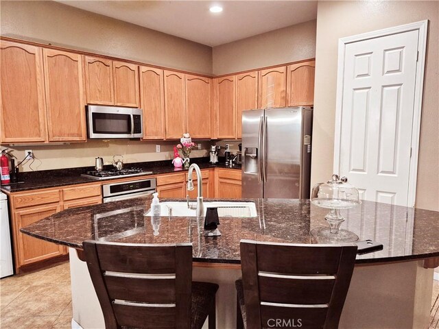 kitchen with stainless steel appliances, dark stone counters, light brown cabinetry, an island with sink, and a kitchen bar