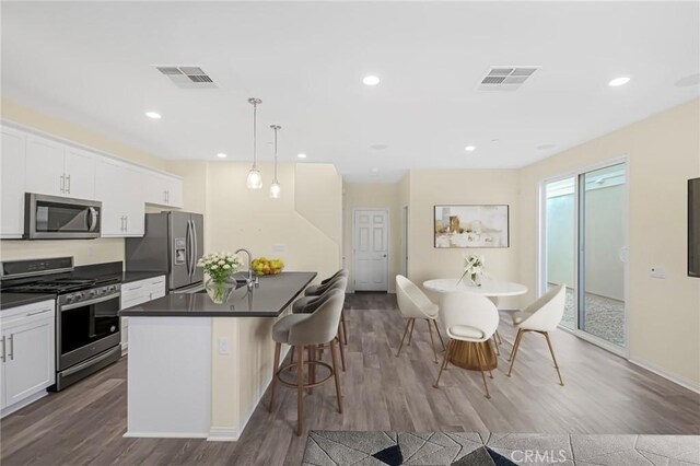 kitchen with a kitchen island with sink, white cabinets, hanging light fixtures, appliances with stainless steel finishes, and dark countertops