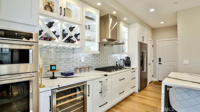 kitchen with beverage cooler, wall chimney exhaust hood, glass insert cabinets, appliances with stainless steel finishes, and white cabinetry