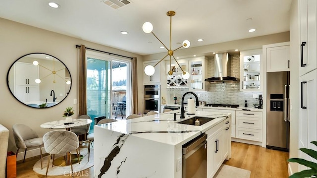 kitchen with decorative light fixtures, a sink, appliances with stainless steel finishes, wall chimney exhaust hood, and a center island with sink