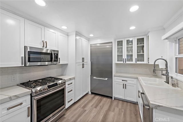 kitchen with light stone counters, a sink, white cabinets, appliances with stainless steel finishes, and glass insert cabinets