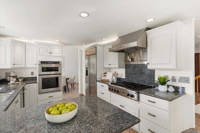 kitchen featuring appliances with stainless steel finishes, white cabinetry, wall chimney range hood, and dark stone countertops