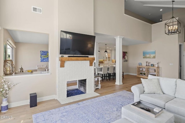 living room featuring visible vents, light wood-style floors, ornate columns, a high ceiling, and a brick fireplace