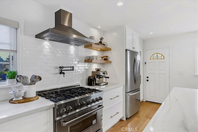 kitchen with wall chimney exhaust hood, light wood-style flooring, appliances with stainless steel finishes, light stone counters, and white cabinetry