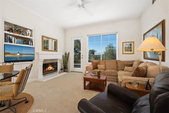 living room with ceiling fan, built in shelves, light colored carpet, ornamental molding, and a glass covered fireplace