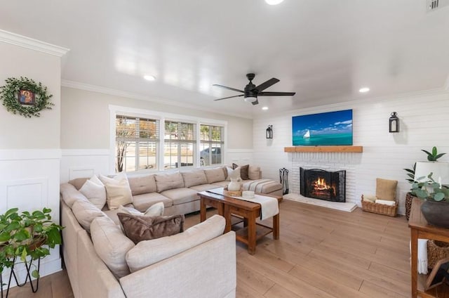 living room featuring light wood finished floors, a wainscoted wall, ceiling fan, crown molding, and a brick fireplace