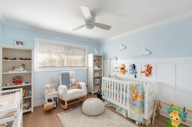 bedroom with a crib, a wainscoted wall, ceiling fan, ornamental molding, and wood finished floors