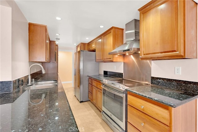 kitchen featuring brown cabinets, appliances with stainless steel finishes, a sink, wall chimney range hood, and dark stone counters