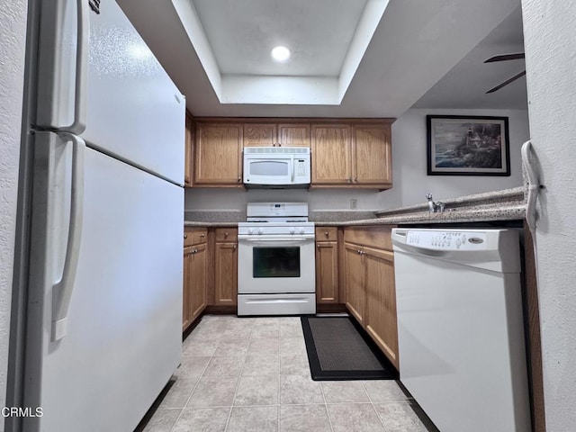 kitchen featuring white appliances, brown cabinetry, dark countertops, a tray ceiling, and light tile patterned flooring