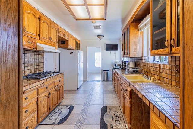 kitchen featuring glass insert cabinets, under cabinet range hood, tile countertops, freestanding refrigerator, and a sink