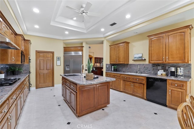 kitchen featuring a sink, brown cabinetry, black appliances, a tray ceiling, and an island with sink