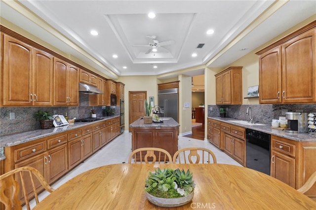 kitchen with stone countertops, a kitchen island with sink, a tray ceiling, black appliances, and a sink