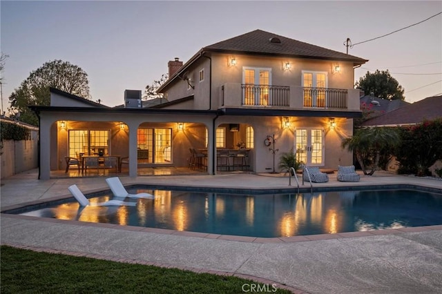 back of property at dusk with a chimney, french doors, a balcony, and a patio