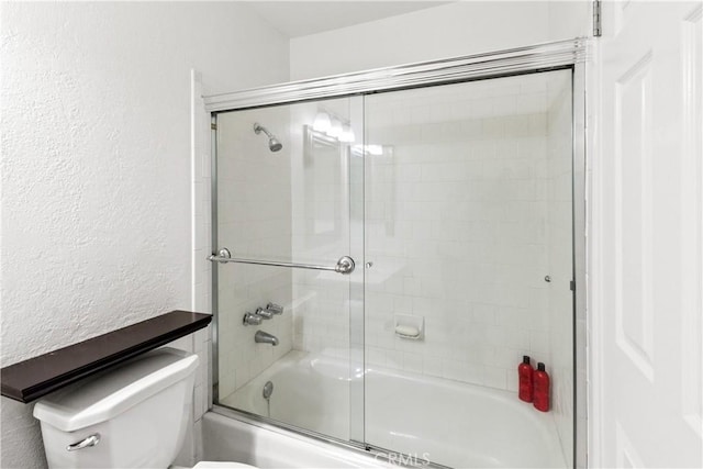 bathroom featuring toilet, combined bath / shower with glass door, and a textured wall