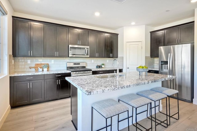 kitchen with light wood-style flooring, appliances with stainless steel finishes, a kitchen island with sink, a sink, and light stone countertops