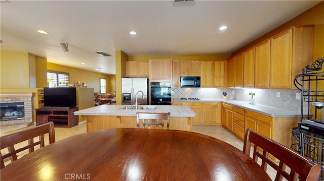 kitchen featuring tile counters, an island with sink, light brown cabinetry, black appliances, and a sink