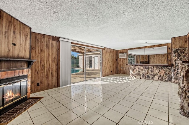 unfurnished living room featuring light tile patterned floors, a textured ceiling, a wall unit AC, wooden walls, and a glass covered fireplace