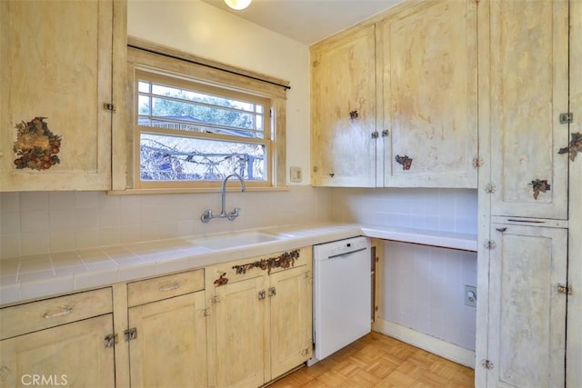 kitchen with tile countertops, backsplash, light brown cabinetry, white dishwasher, and a sink