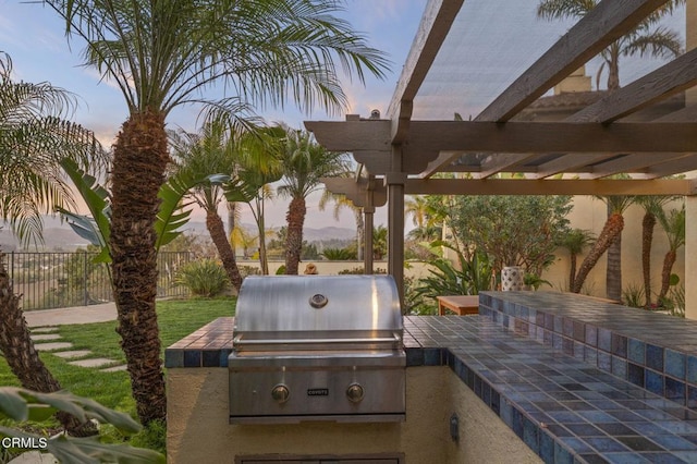 patio terrace at dusk with a grill, fence, an outdoor kitchen, and a pergola