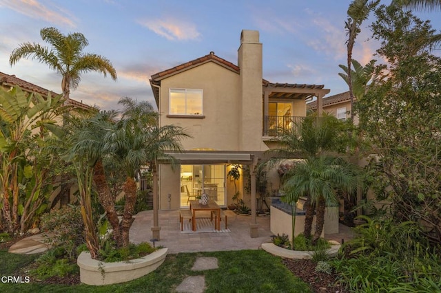 back of house at dusk featuring a balcony, a patio area, a chimney, and stucco siding