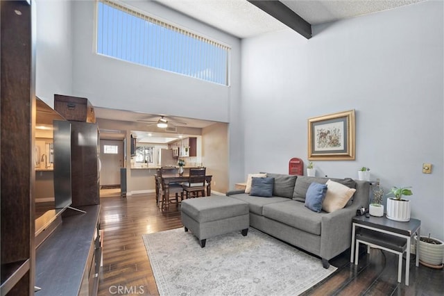 living area with baseboards, a towering ceiling, dark wood-style floors, ceiling fan, and beamed ceiling